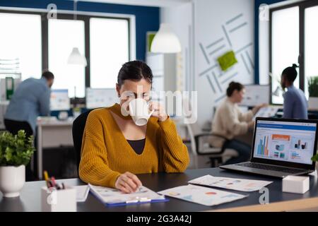 Brand manager businesswoman drinking a cup of coffee analysing financial start up charts. Executive entrepreneur, manager leader sitting working on projects with diverse teamworkers. Stock Photo