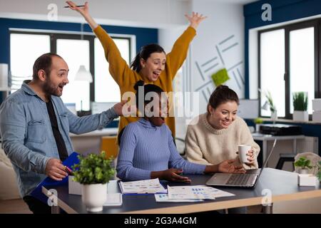 Diverse team celebrating triumph achievement of partnership with hands up reading good news on laptop. Multiethnic business team with laptop and papers excited about project. Stock Photo