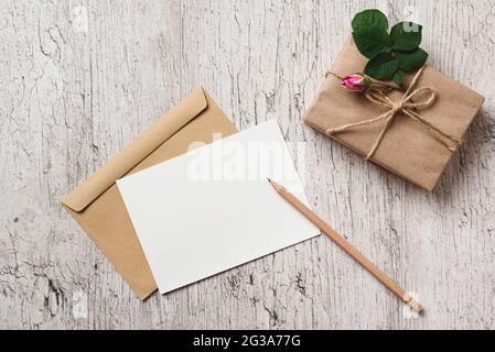 Mockup blank white greeting card or invitation, gift box wrapped with kraft paper and pink rose bud on gray table. Blank sheet of paper for text Stock Photo
