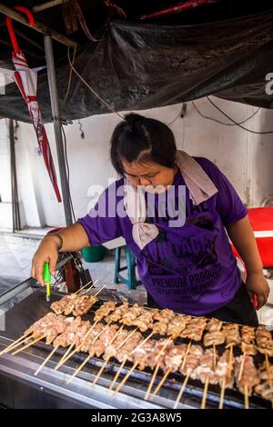 A Thai street food vendor cooks her chicken satay sticks on a grill at her kiosk in the Bang Rak neighborhood of Bangkok. Stock Photo