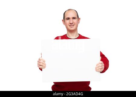 A bald young man dressed in a red sweater is holding with his hands a blank poster in front of himself placed horizontally. The background is white. Stock Photo