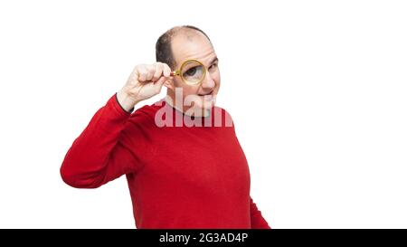 A bald young man wearing a red sweater is looking forward through a magnifying glass and his right eye is very large. The background is white. Stock Photo