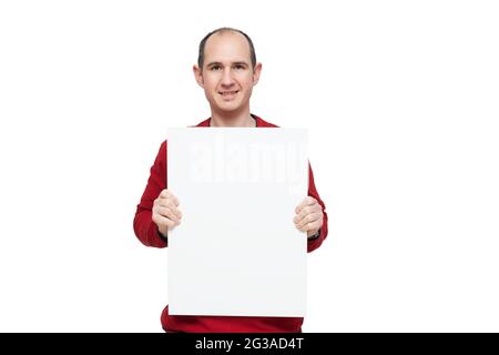 A bald young man dressed in a red sweater is holding with his hands a blank poster in front of himself placed vertically. The background is white. Stock Photo