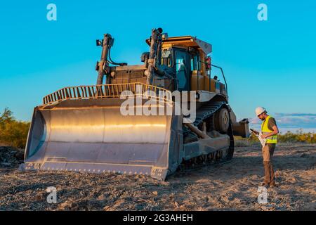 Hipster construction manager with a beard, safety helmet and reflective vest looks at his smartphone while inspecting work machinery on a construction Stock Photo