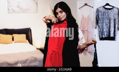 young woman in pink silk peignoir trying on dresses on hangers in bedroom Stock Photo