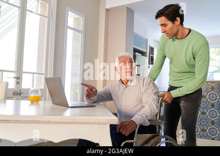 Young man helping senior in wheelchair on laptop computer with social media communication Stock Photo