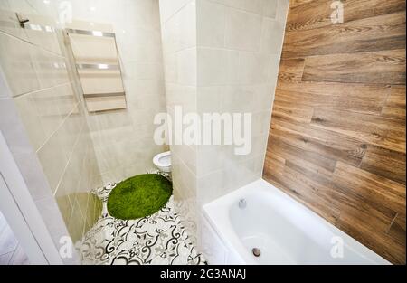 View of left part of small light bathroom with modern renovation. White tiles on the walls combining with wooden elements, ceramic plumbing and green bath mat. Stock Photo