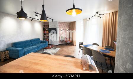Wide angle view of minimalist living room in beige, gray and blue tones with window, curtains, parquet, sofa, armchair, carpet, tables, rack, TV set. Pendant lamps in loft style in different versions. Stock Photo