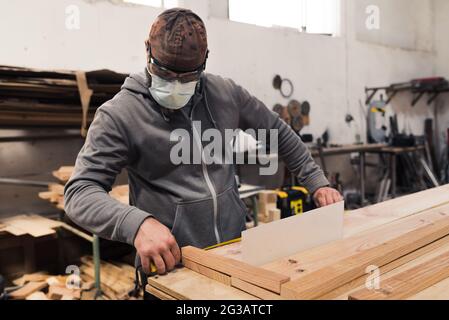 Carpenter with mask and cap taking measurements on a wood with a tape measure Stock Photo