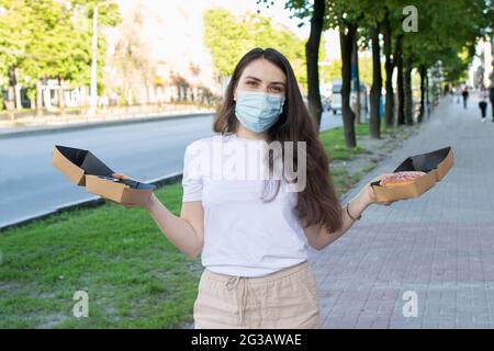 A masked woman deliverer of food during a pandemic and quarantine keeps food to order in cardboard boxes. Street food and coronavirus. Stock Photo