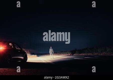 A mysterious lone figure next to a car, parked on the side of the road,  on a summers night. Standing in the middle of a road looking up at the night Stock Photo