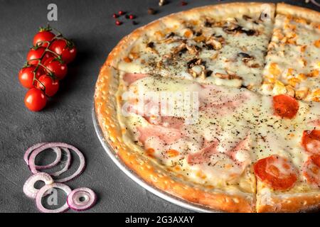 Italian pizza four seasons with cheese, ham, mushrooms, tomato, onion, pepperoni sausage on a gray table, close-up Stock Photo