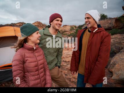 Cheerful young male and female friends in winter clothing laughing during camping on mountain hill Stock Photo