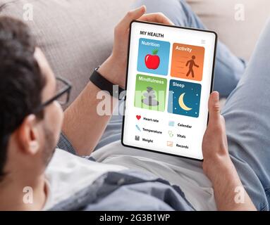 Young man with glasses enjoying brand new health app on digital tablet while reclining on couch at home, over shoulder shot, closeup. Healthcare mobil Stock Photo