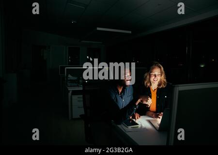Two diverse happy colleagues working on laptop until night in dark office Stock Photo
