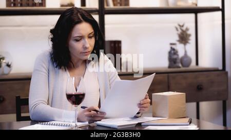 concentrated woman sitting on table with papers and glass of red wine at home Stock Photo