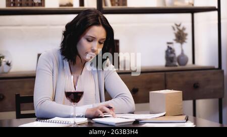 upset woman sitting on table with glass of red wine and counting on calculator at home Stock Photo