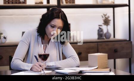 serious woman sitting on table with glass of red wine and writing in notebook at home Stock Photo
