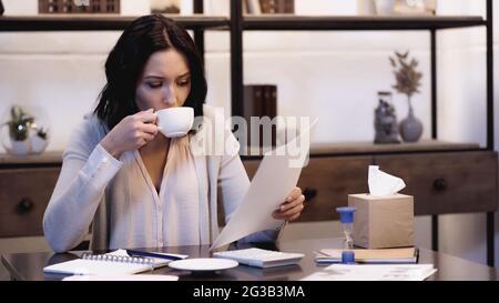 brunette woman sitting on table, drinking coffee and looking at documents at home Stock Photo