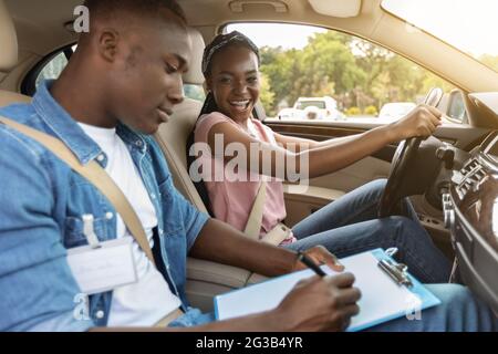 Young black man instructor examinating happy lady student, taking notes at chart while sitting by cheerful african american woman driving auto, side v Stock Photo