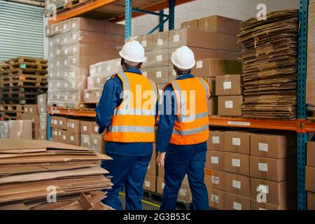 Rear view of two male workers in safety workwear walking near the shelves with stack of goods in warehouse Stock Photo