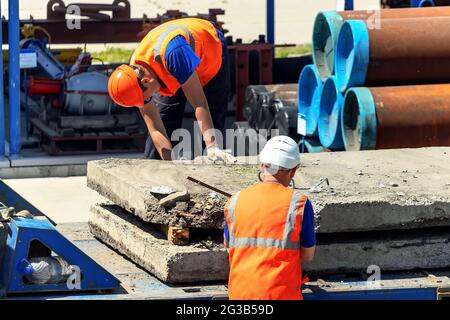 Two slingers unload concrete slabs on the street on a summer day. Workers in construction helmets and vests sling cargo. Work in an open warehouse. Stock Photo