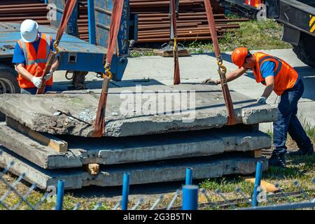 Two slingers unload concrete slabs on the street on a summer day. Workers in construction helmets and vests sling cargo. Work in an open warehouse. Stock Photo