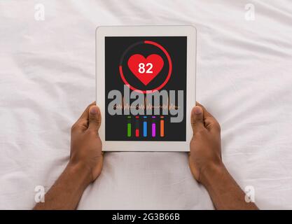 Unrecognizable black man laying in bed and checking his health after waking up, using brand new mobile application on digital tablet, inspecting his h Stock Photo
