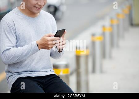 Close-up of smiling young Asian man in gray sweater sitting on metal street pole and checking smartphone Stock Photo
