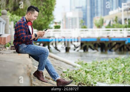 Smiling young Vietnamese man in casual outfit sitting on ground and texting message on smartphone while chatting online Stock Photo
