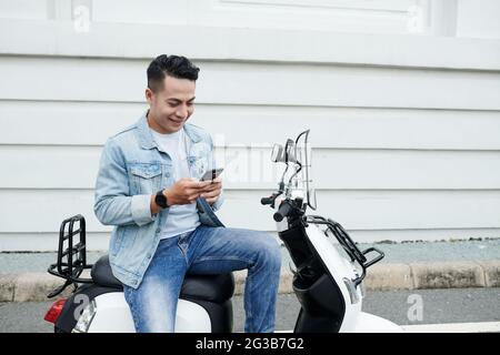 Smiling young Vietnamese moped driver in denim outfit sitting on modern bike and using smartphone while communicating online Stock Photo