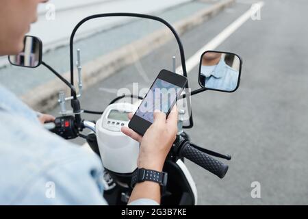 Over shoulder view of man in wristwatch driving moped and using smartphone while navigating with online app Stock Photo