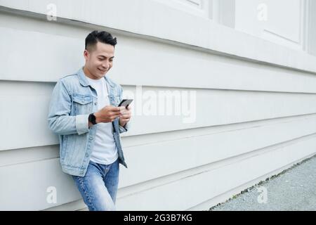 Happy young Asian man in denim outfit standing at white building wall and surfing net on gadget Stock Photo