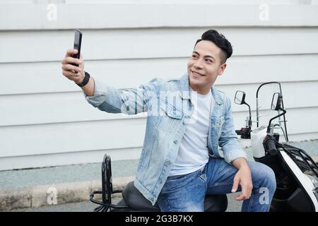 Positive young Vietnamese man in denim clothes sitting on moped and taking selfie on smartphone against street building Stock Photo