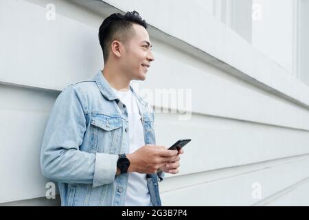 Smiling handsome young Vietnamese man in denim jacket standing with smartphone against building wall Stock Photo
