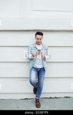 Serious fashionable young Vietnamese man in denim clothes holding foot on wall and using modern phone Stock Photo