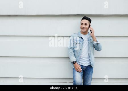 Positive young Asian man in casual outfit leaning on building wall and having phone conversation Stock Photo