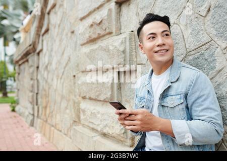 Smiling handsome young Asian man in denim jacket standing at old wall and using gadget while looking aside Stock Photo