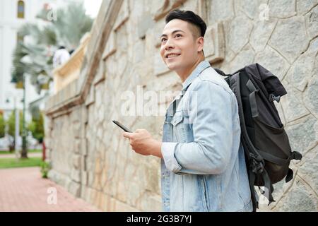 Positive young Asian man with black backpack using navigation app on phone and walking in old city while travelling alone Stock Photo