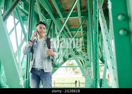 Smiling young Vietnamese guy with satchel chatting by mobile phone while walking under bridge with green metal girders Stock Photo