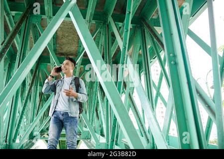 Positive young Vietnamese tourist in denim outfit standing under bridge with green metal girders and gesturing hand while chatting by phone Stock Photo