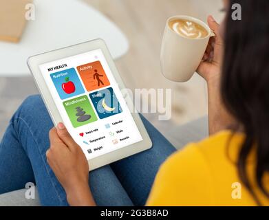 Young black woman with cup of coffee sitting on sofa and using digital tablet at home, checking her health condition via brand new mobile application, Stock Photo