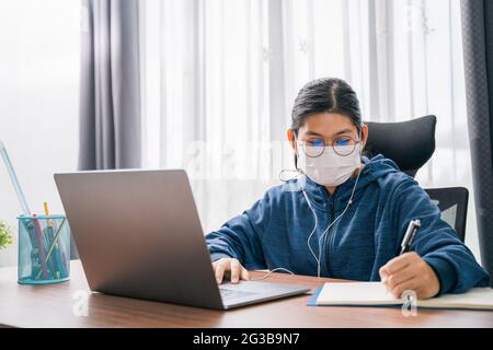 Asian young girl wear a mask student with glasses headphones girl study happy writing note on a book looking video conference laptop computer online i Stock Photo
