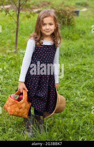 Cute Russian Six Year Old Girl Hold Basket With Fresh Ripe Red Apple And Wicker Hat In Garden. Harvesting Fresh Apples. Stock Photo