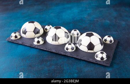 Halves of chocolate football balls of different sizes on a black plate on the blue table. A chocolate dessert concept for a fan of European football Stock Photo