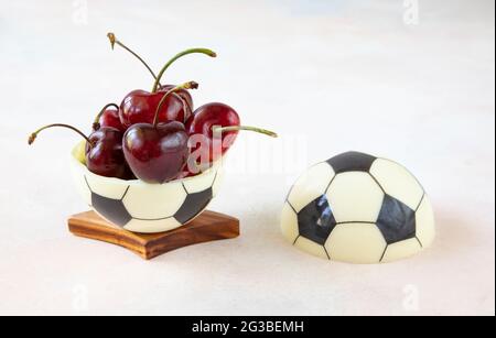 A sweet dessert for a fan of european football - a chocolate black and white ball made of two halves with ripe red cherries with sprigs on the table Stock Photo
