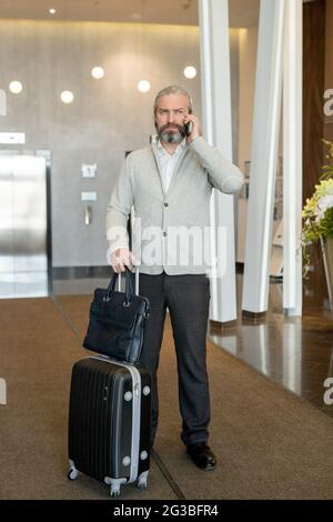 Mature businessman with suitcase talking on mobile phone in hotel lounge Stock Photo