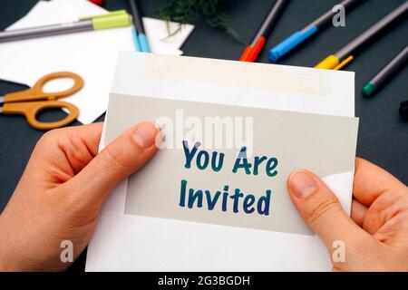 Woman hands taking out gray paper with words You Are Invited from envelope. Office supplies on background. Stock Photo