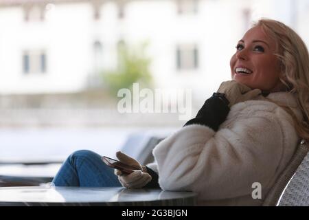 Joyful young woman relaxing outdoors on a patio in winter seated at a table with her mobile in hand looking up with a vivacious smile Stock Photo