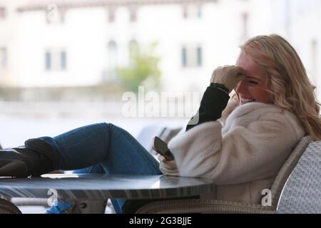 Natural portrait of an amused vivacious young woman laughing happily as she sits on an outdoor patio in winter using her mobile phone Stock Photo